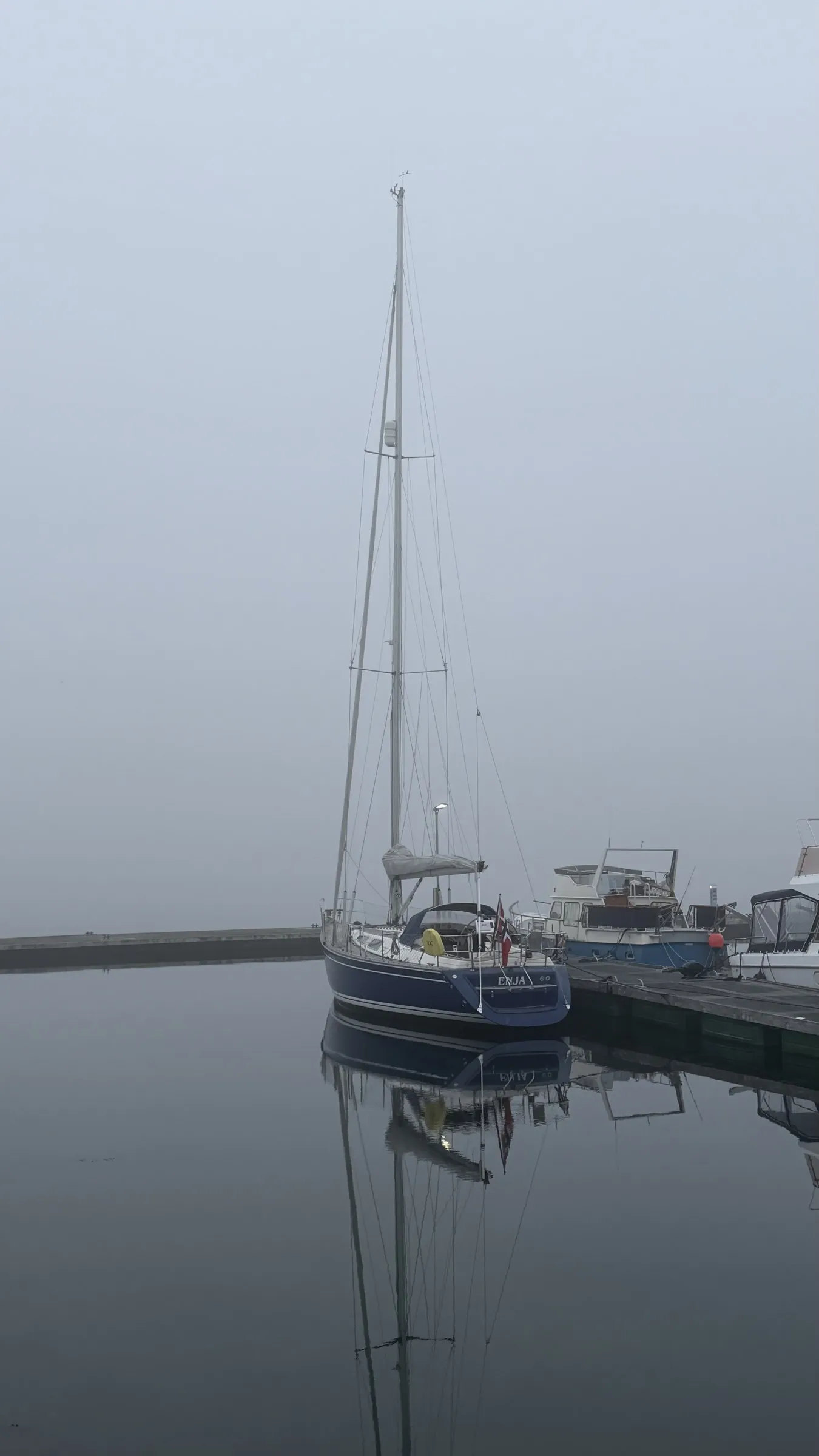 Enja alongside in flat calm fog, her mast and hull mirrored in the water