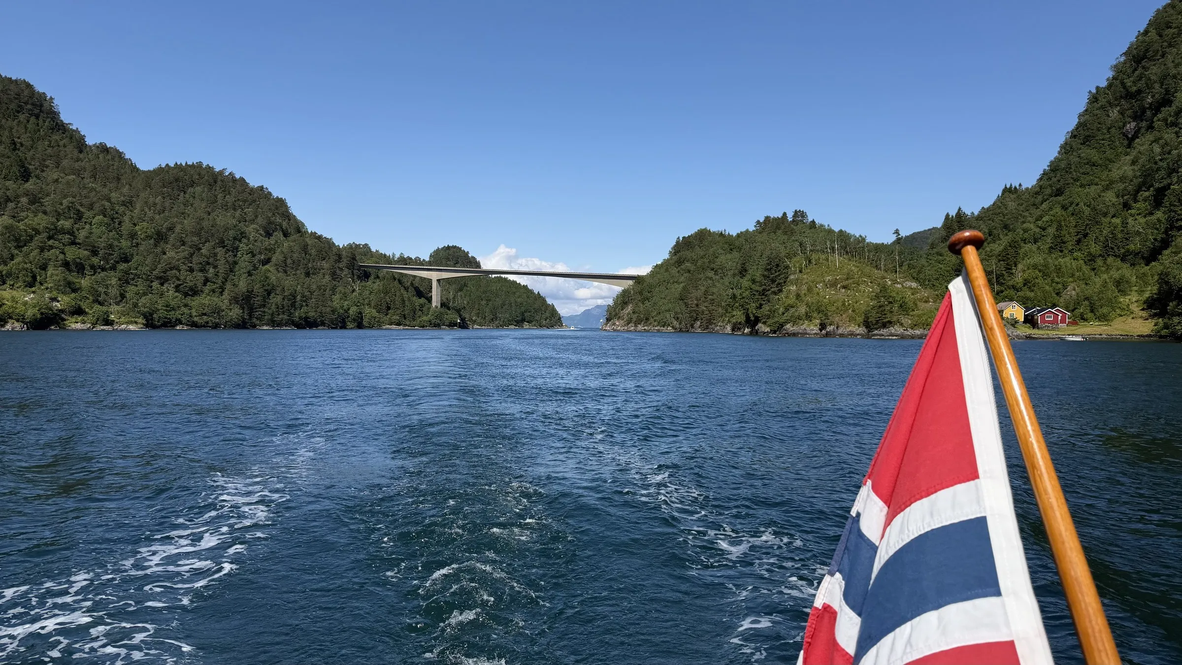 A narrow sunlit fjord with wooded sides and a high bridge ahead, the ensign in the foreground and the wake astern