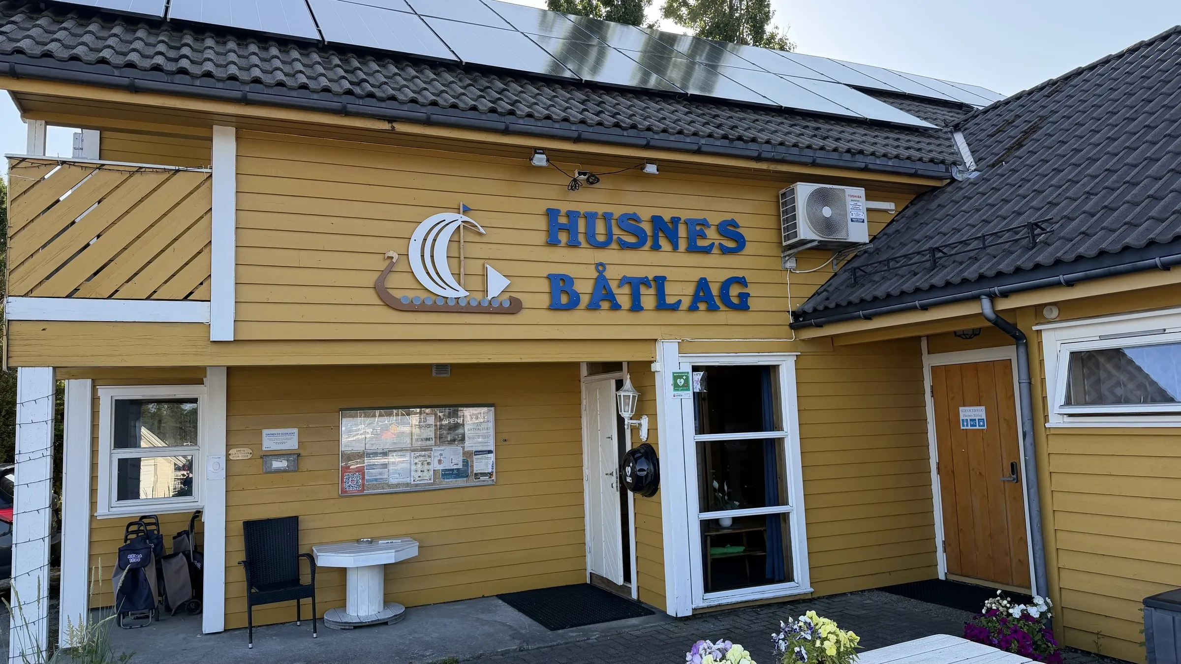 A yellow clubhouse in evening sun with a blue sign reading Husnes Båtlag above the door